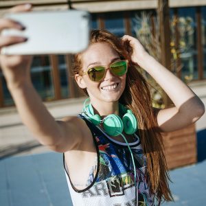 Smiling young woman wearing mirrored sunglasses taking selfie with smartphone