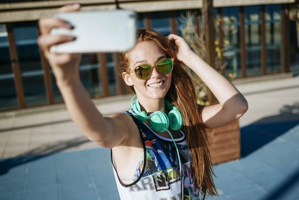 Smiling young woman wearing mirrored sunglasses taking selfie with smartphone