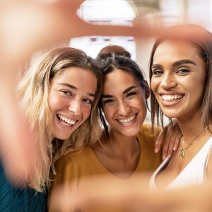 Three multiracial female friends taking selfie and making hand frame gesture