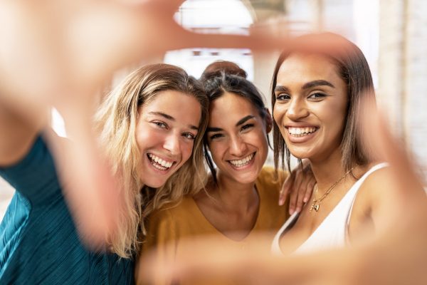 Three multiracial female friends taking selfie and making hand frame gesture